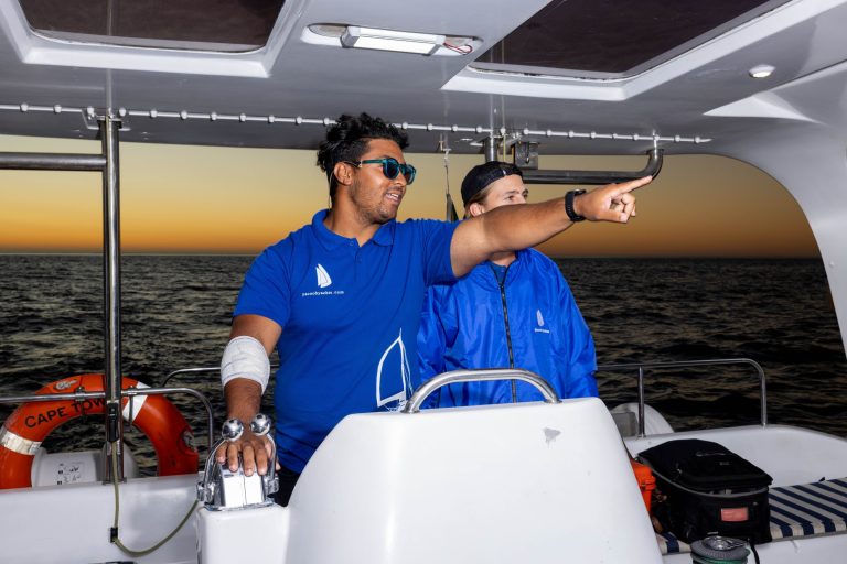 Two people in blue jackets on a boat, one pointing at sea during sunset.