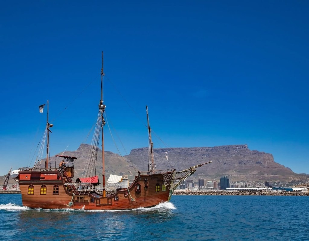 Wooden pirate ship sailing with cityscape and mountains in background.