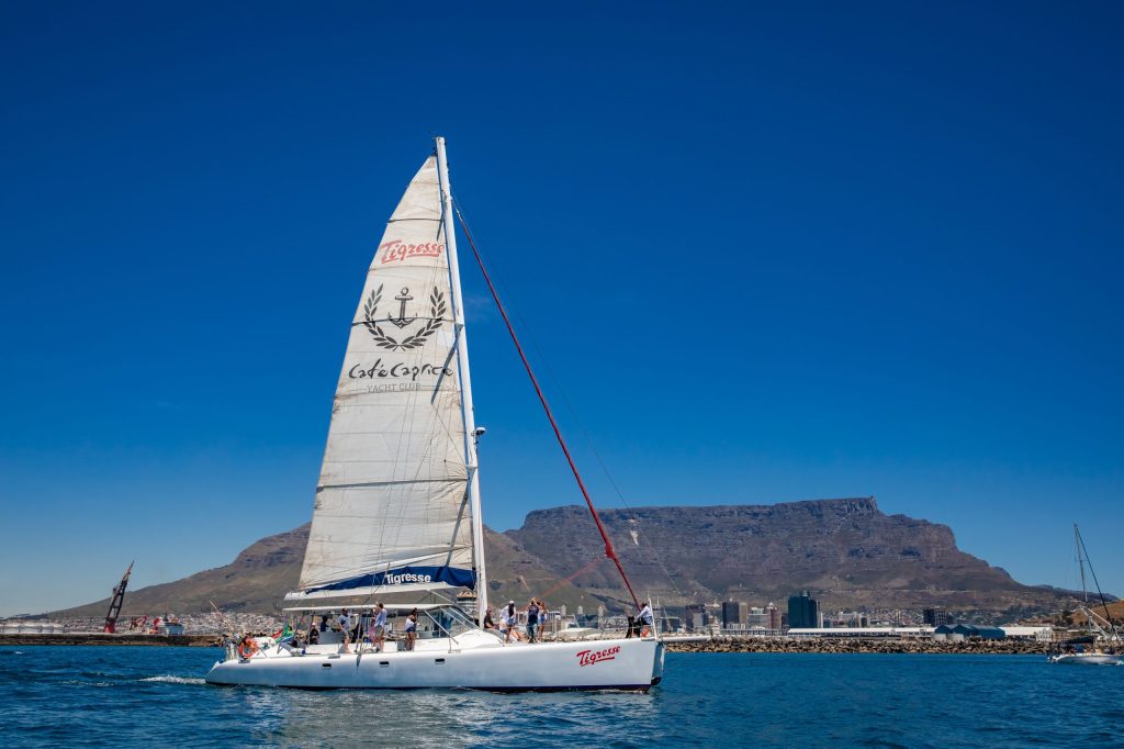 White sailboat with people on a blue sea, distant mountains in background.