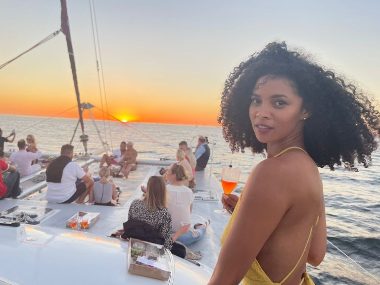 Woman holding a drink on a boat at sunset with people in the background.