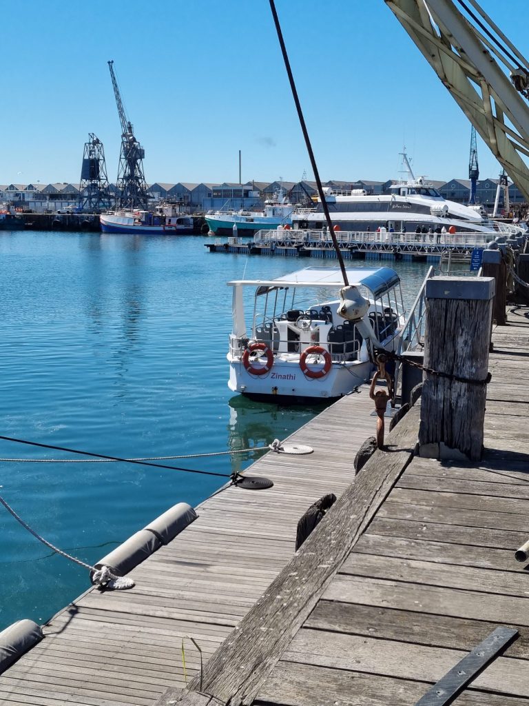 Boat docked at a wooden pier with cranes and buildings in the background on a sunny day.