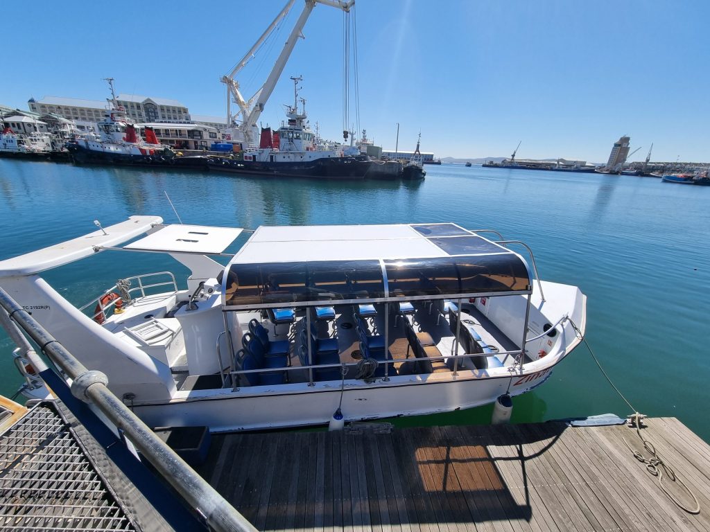 Small tour boat docked in sunny harbor, cranes and ships in background.