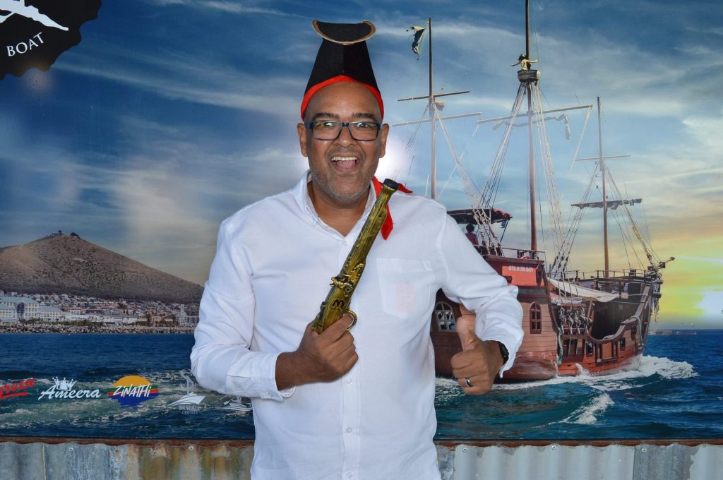 Smiling person in pirate hat gives thumbs-up near ship mural.