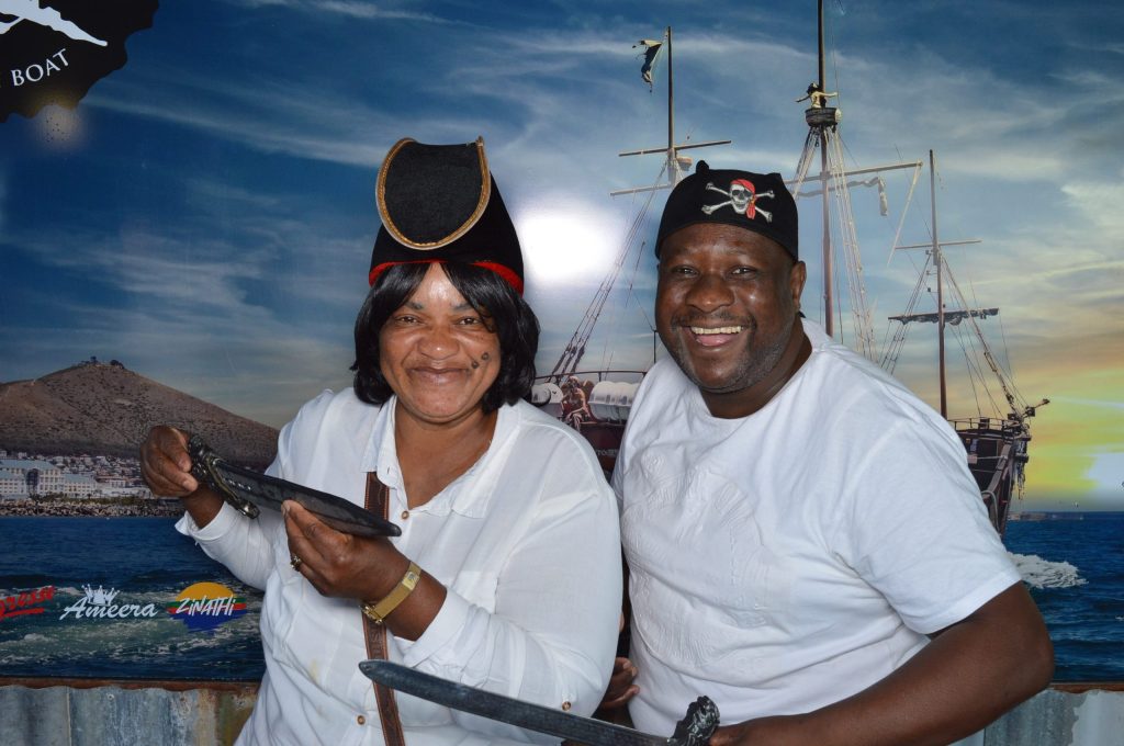 Two smiling people in pirate hats holding swords with a ship backdrop.