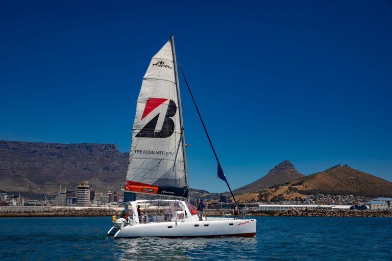 Sailing catamaran on water with cityscape and mountains in background.