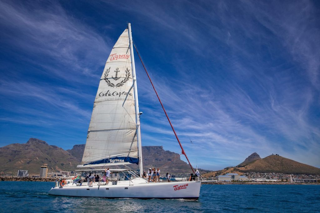 White sailboat with people on board on a sunny day with mountains in the background.