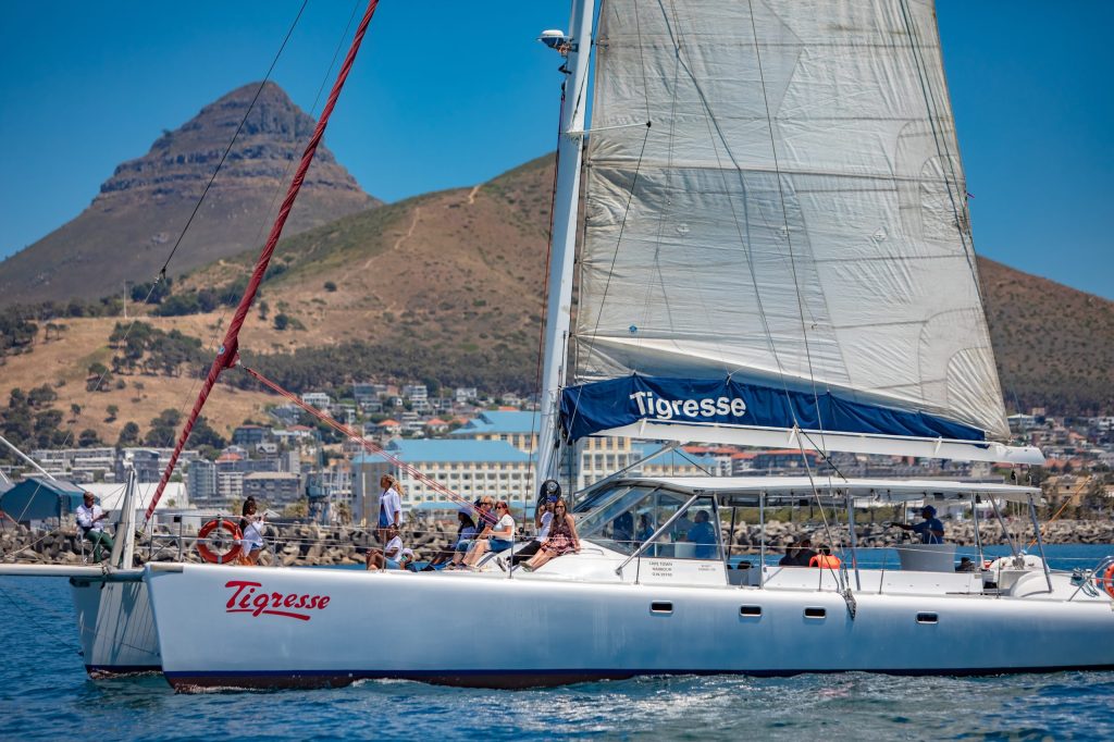 Sailboat named Tigresse with people on board, sailing near a mountain coastline.