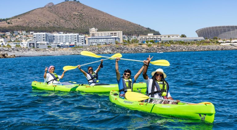 Three people kayaking on a sunny day with mountains and buildings in the background.