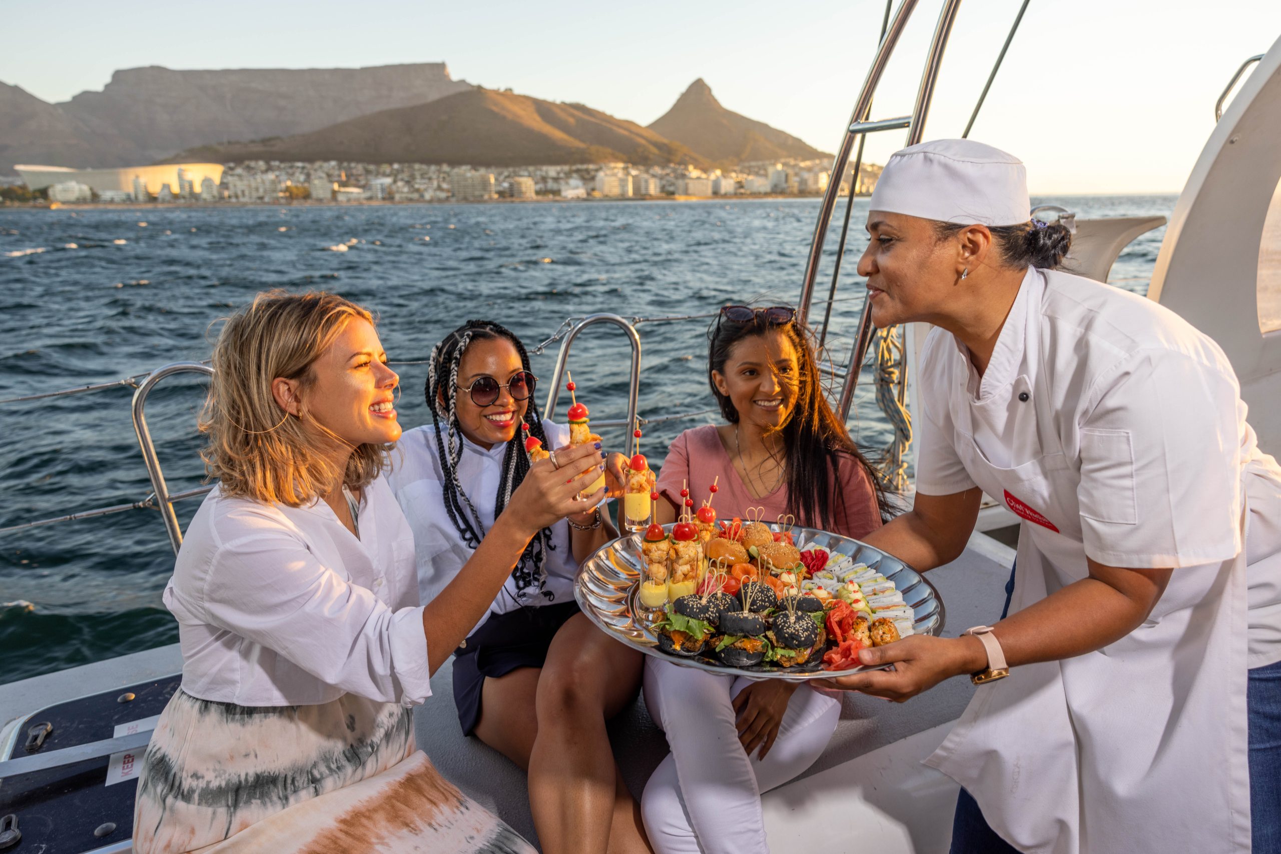 Group of women on a boat enjoying refreshments served by a chef.