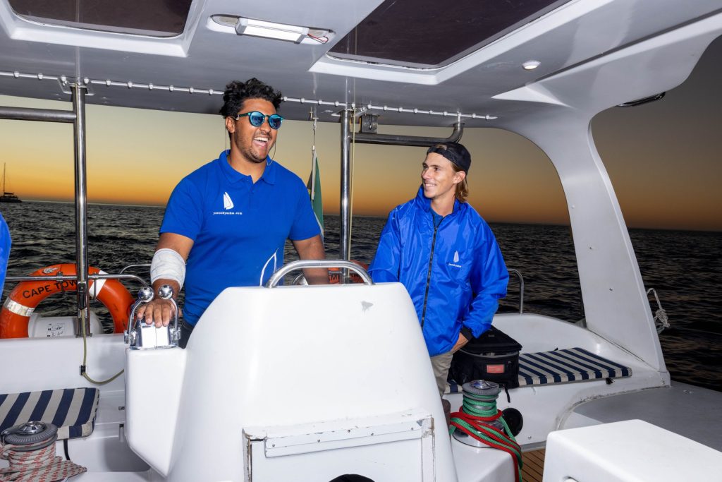 Two people on a boat at sunset, one steering, both in blue shirts and jackets, ocean in the background.