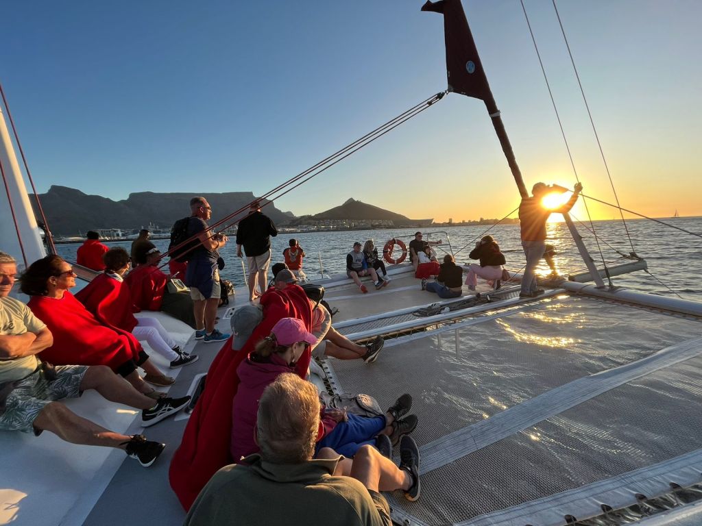 People relaxing on a catamaran at sunset, with mountains in the background.