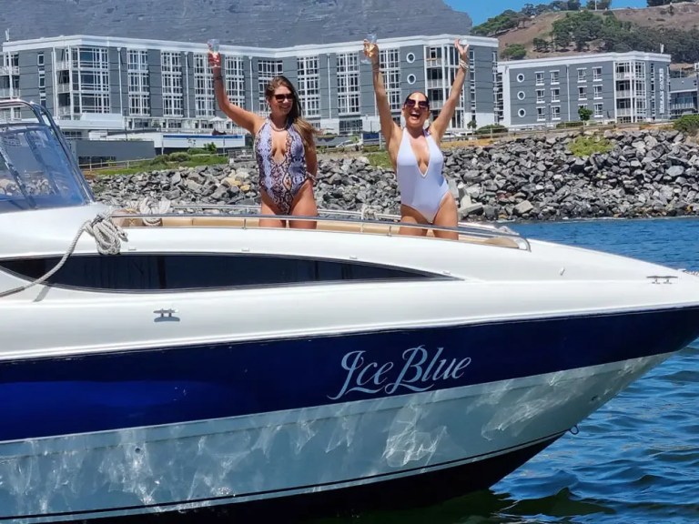 Two people in swimsuits on a boat named 'Ice Blue', holding drinks, with buildings and a rocky shore in the background.