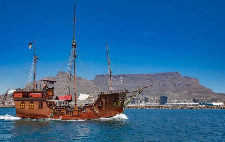 Wooden ship sailing with mountains and cityscape in the background.